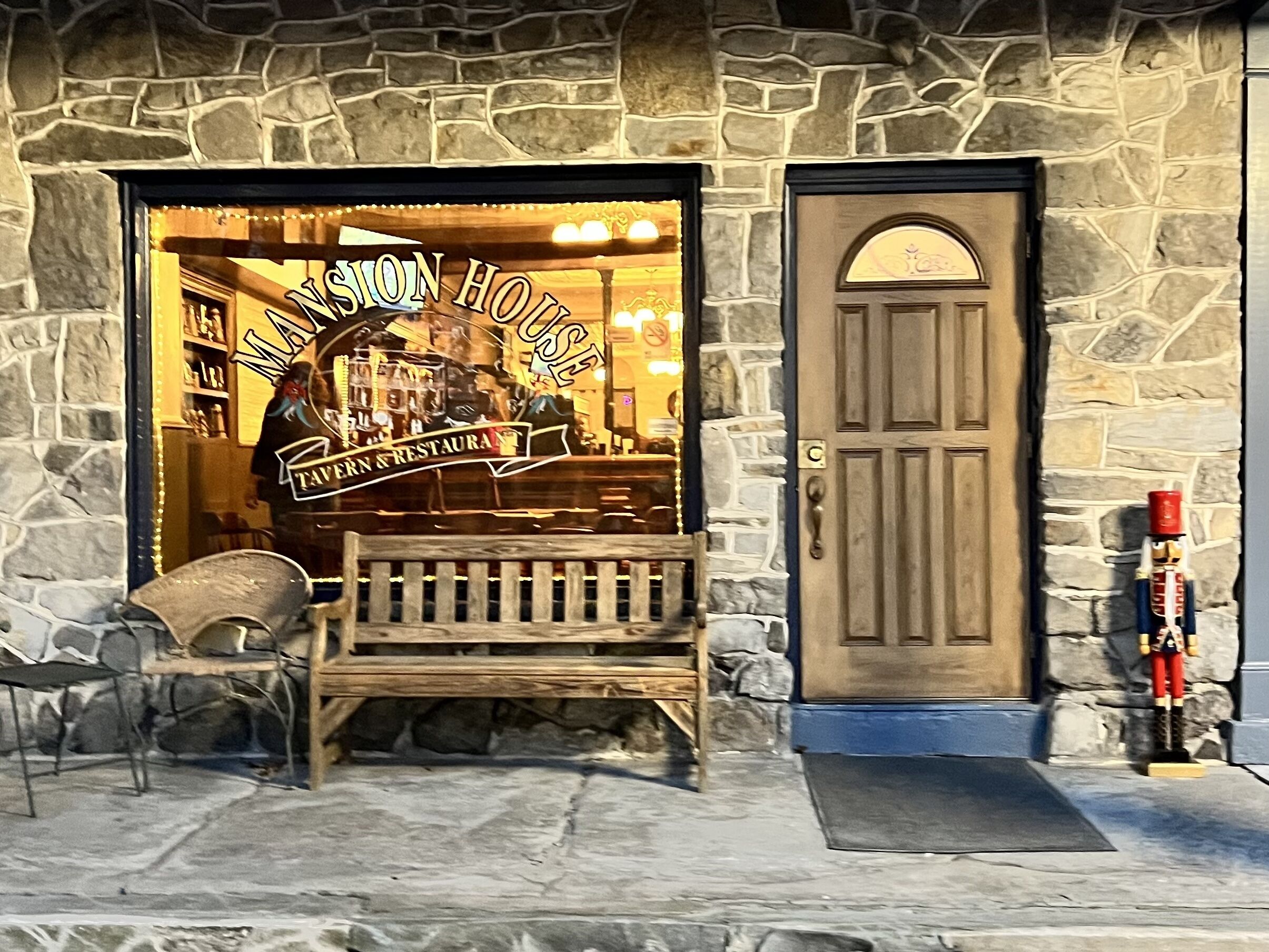 A stone building with a wooden bench outside, a door, and a sign that reads "Mission House Tavern & Restaurant".