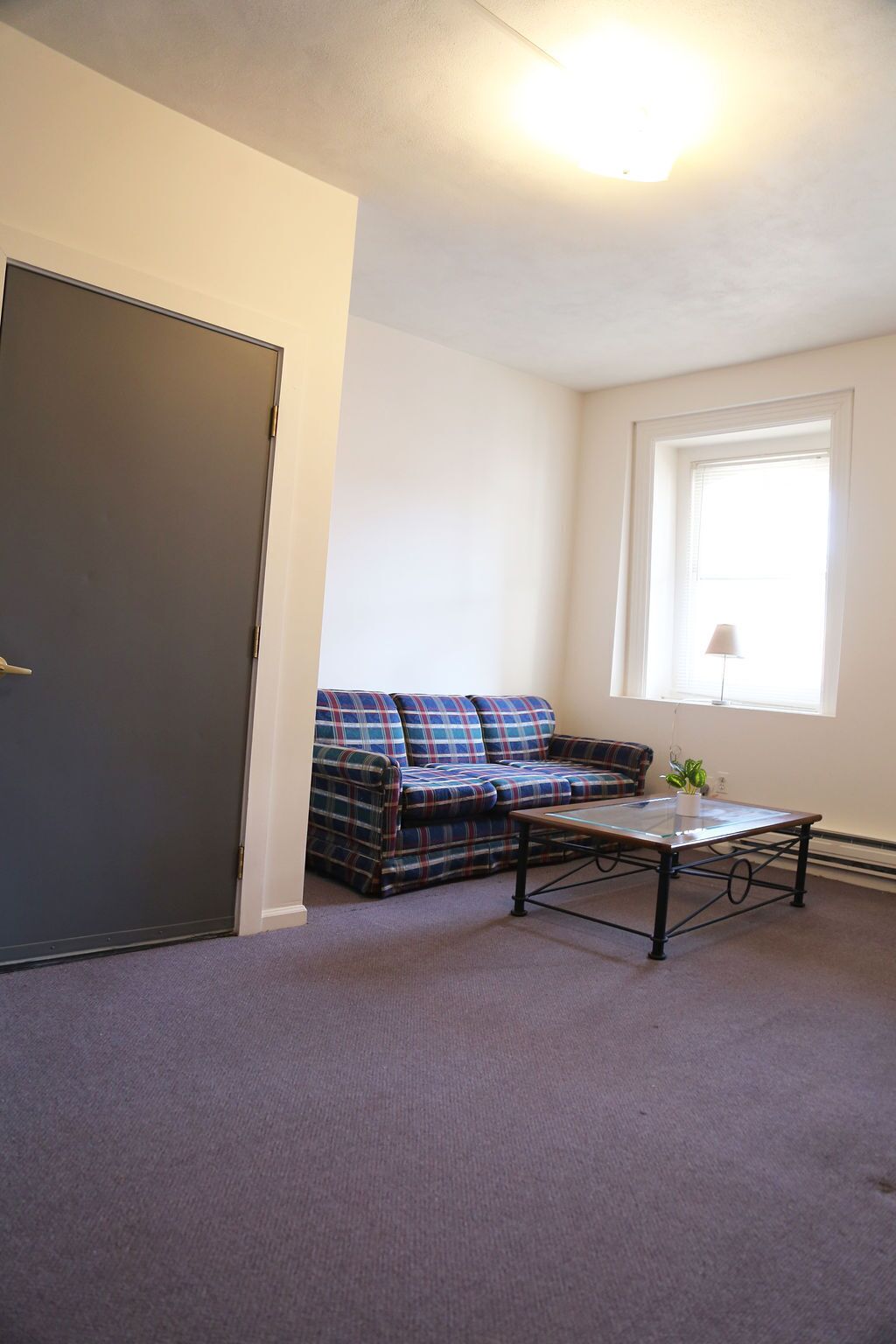 Una habitación con un sofá a cuadros, una mesa de centro de vidrio, una puerta cerrada y una ventana con persianas.