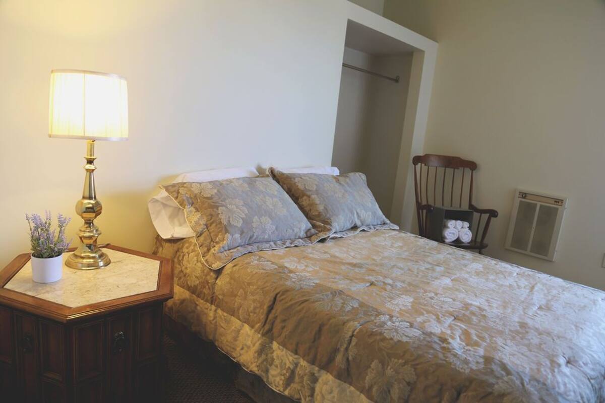 A bedroom with a bed, a nightstand with a lamp, a chair, and a radiator.