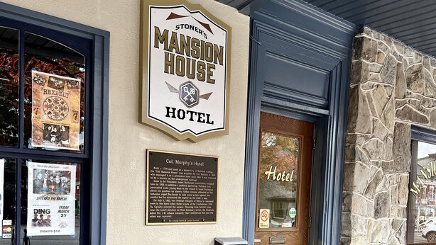 Entrada de un hotel con un letrero que dice "Stoner's Mansion House Hotel". Hay sillones de mimbre afuera y una placa con información histórica.