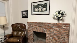 Una habitación con chimenea de ladrillo, un sillón de cuero, un cuadro con cisnes y un jarrón con flores blancas.