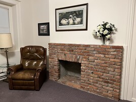 Una habitación con chimenea de ladrillo, un sillón de cuero, un cuadro con cisnes y un jarrón con flores blancas.