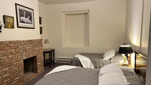 Habitación de hotel con chimenea de ladrillo, dos camas con ropa de cama gris, una mesita de noche con lámpara y cuadros enmarcados en la pared.