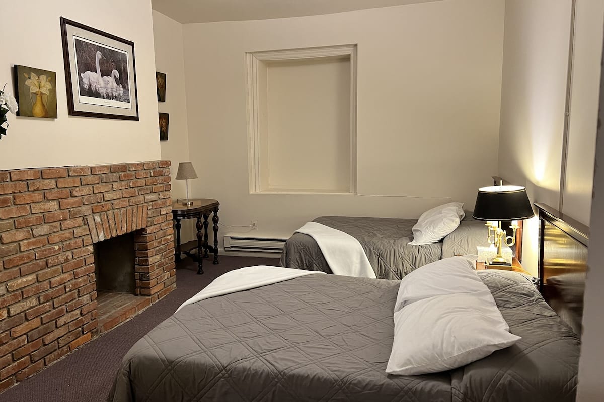 A hotel room with a brick fireplace, two beds with grey bedding, a small table with a lamp, and framed pictures on the wall.