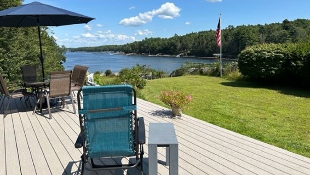A deck with outdoor furniture, a flag, and a view of a lake. - Spectacular sunrises,cozy vintage home on the water in Wiscasset, ME-Adults only (Wiscasset)