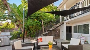 Ein möblierter Patio-Bereich im Freien mit Korbmöbeln, einem Grill und einer Treppe, die zu einem Balkon führt.