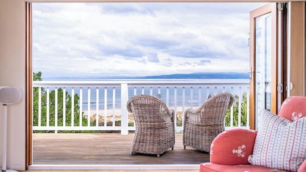 A balcony with wicker chairs and a red sofa, overlooking a beach and ocean. - Hamptons Hideaway - Freycinet Holiday Houses (Coles Bay)
