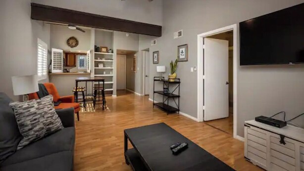 Una sala moderna con sofá, mesa de centro y televisor. Se observa un área de cocina al fondo con una mesa de comedor y sillas.