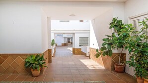 Un couloir au sol carrelé, agrémenté de plantes en pot, d’où l’on aperçoit un bâtiment doté d’une porte et de fenêtres.