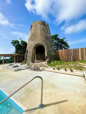 Uma construção de pedra com entrada em arco, uma piscina com grade de metal e área externa com cadeiras e mesas.