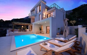 A modern two-story house with a swimming pool, outdoor seating, and a mountain backdrop.