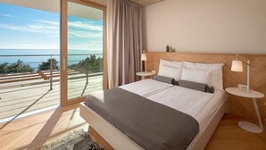 A modern bedroom with a large bed, two bedside tables, a view of the sea, and a balcony.