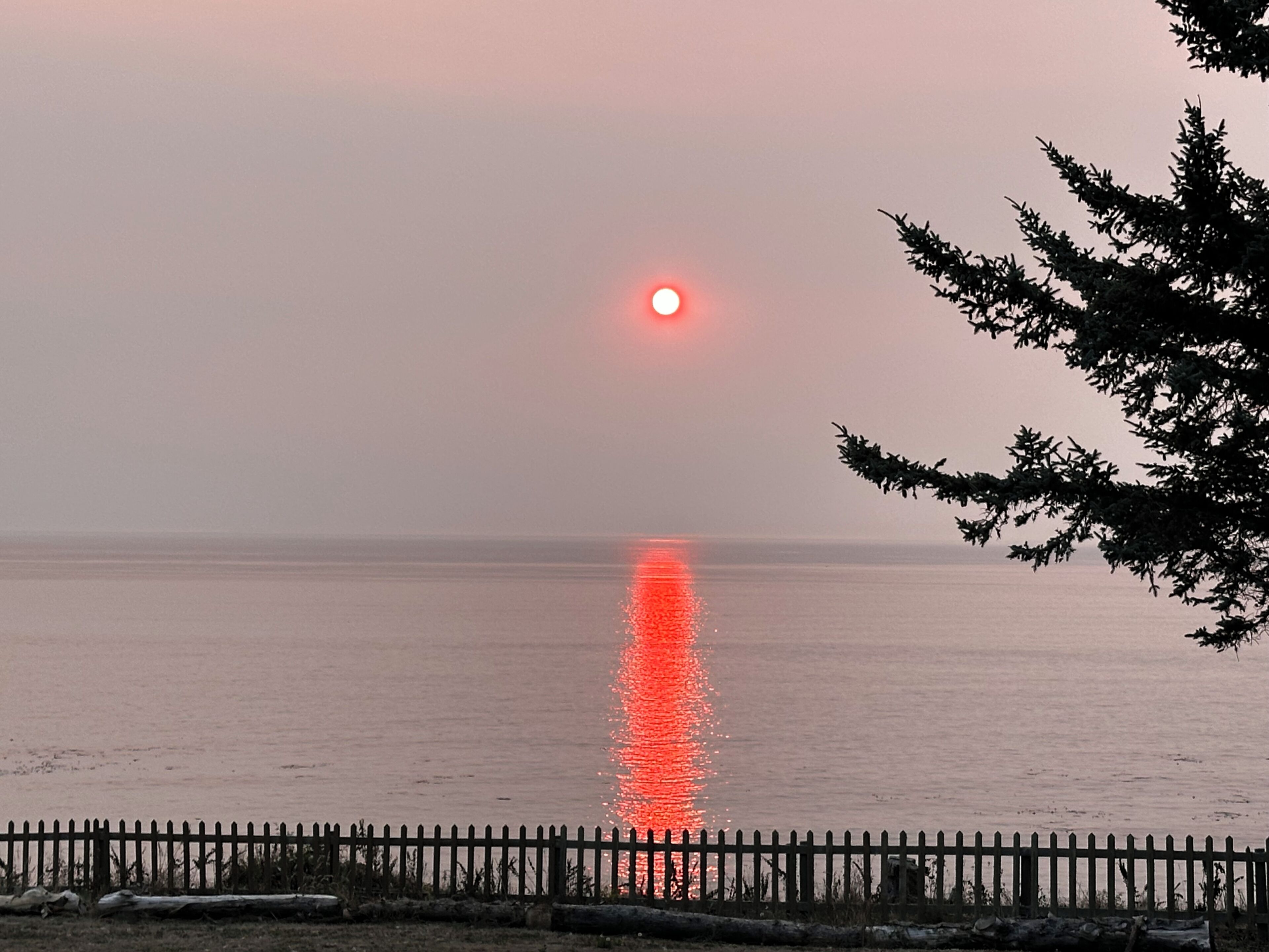 Un coucher de soleil sur une étendue d’eau calme, avec une clôture en bois au premier plan.
