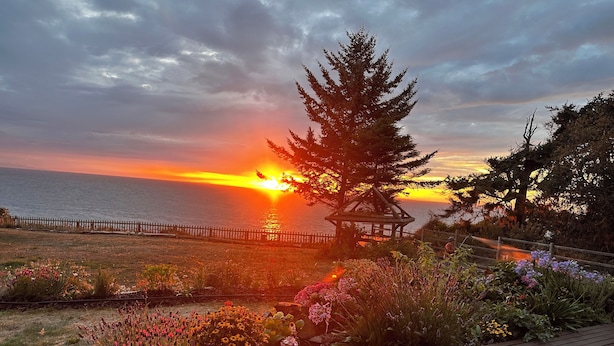 A wooden deck with a gazebo, a grassy area with flowers, and a view of the sunset over the ocean. - Home Away From Home… for you and for your pets! (Oak Harbor)