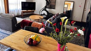 A living room with a sofa, a coffee table, a TV, a wall-mounted exercise bike, and a dining area with a fruit bowl and flowers. - Home Away From Home… for you and for your pets! (Oak Harbor)
