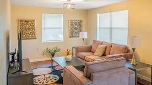 A living room with a sofa, coffee table, and decorative items. There are two windows with blinds, a floor lamp, and a TV stand. - Comfort Living Near LRMC, Medical Row
 (Lakeland)