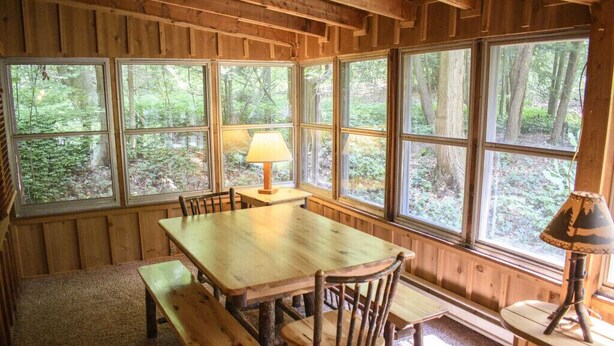 Une cabane en bois dotée d’une grande table à manger et de bancs, entourée de grandes fenêtres qui offrent une vue sur la forêt.