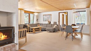 A cozy living room with a fireplace, a sofa, a dining table with chairs, and large windows. - 5 bedroom amazing home in Herning (Herning)