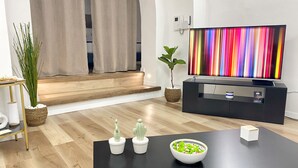 A modern living room with a flat-screen TV, a black coffee table, a bowl of salad, two potted plants, and a staircase leading to an upper level.