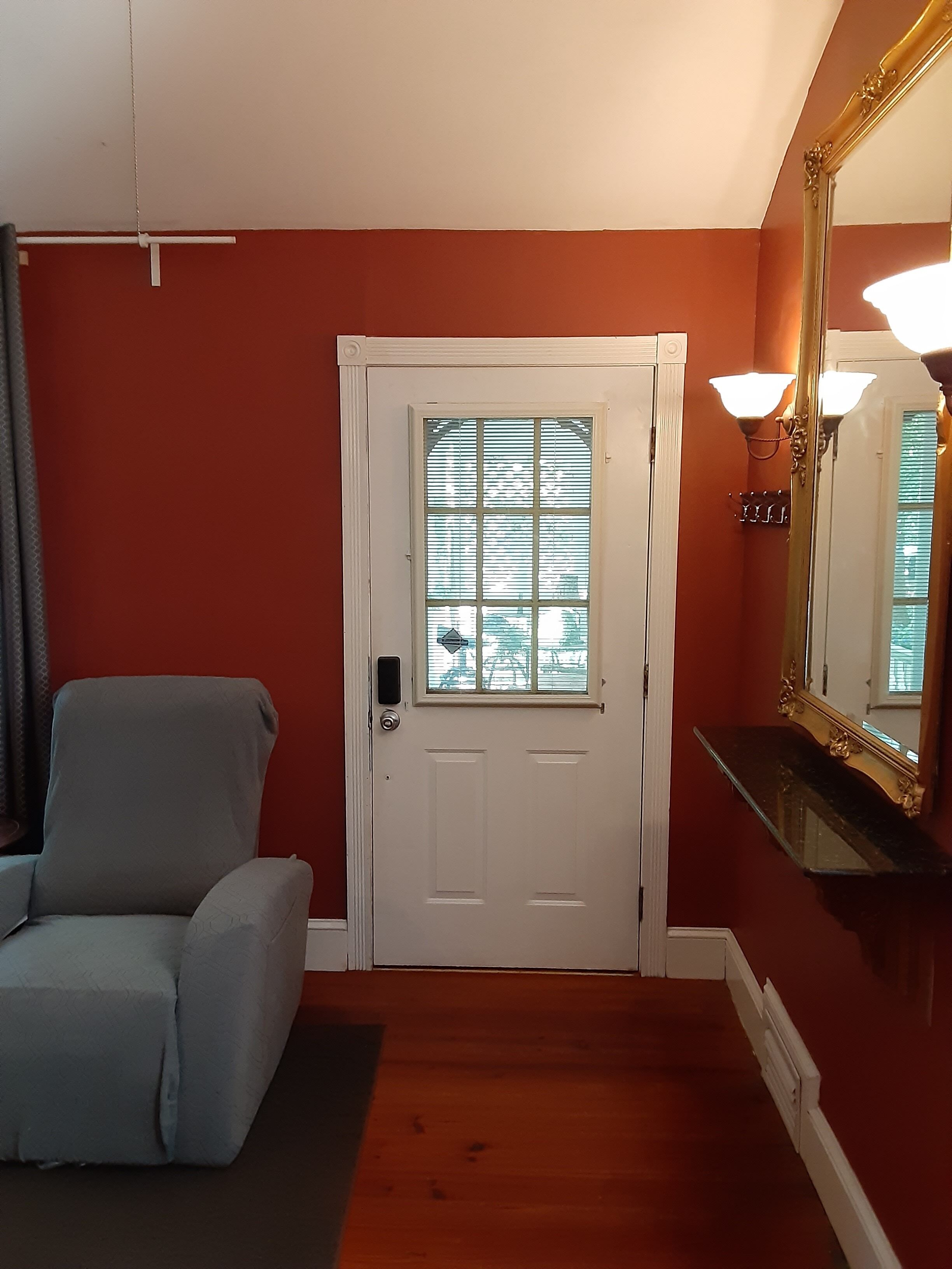 Une pièce avec une porte blanche, un fauteuil gris, un miroir avec un cadre doré et un plancher en bois.