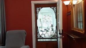 Una habitación con pared roja, un sillón gris, suelo de madera, una puerta blanca con un panel de vidrio decorativo y un espejo con marco dorado.