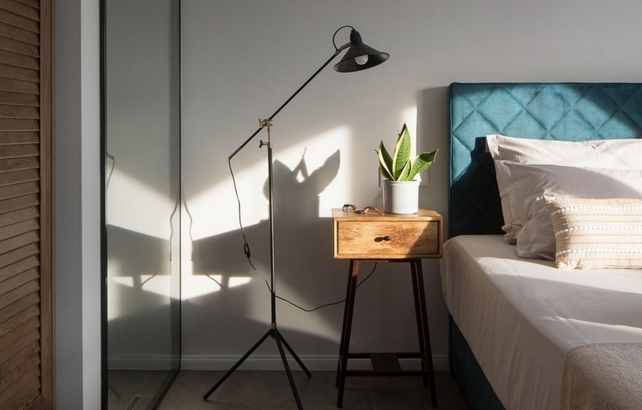Una camera da letto con un letto, un comodino con una pianta, una lampada da terra e una finestra che proietta ombre.