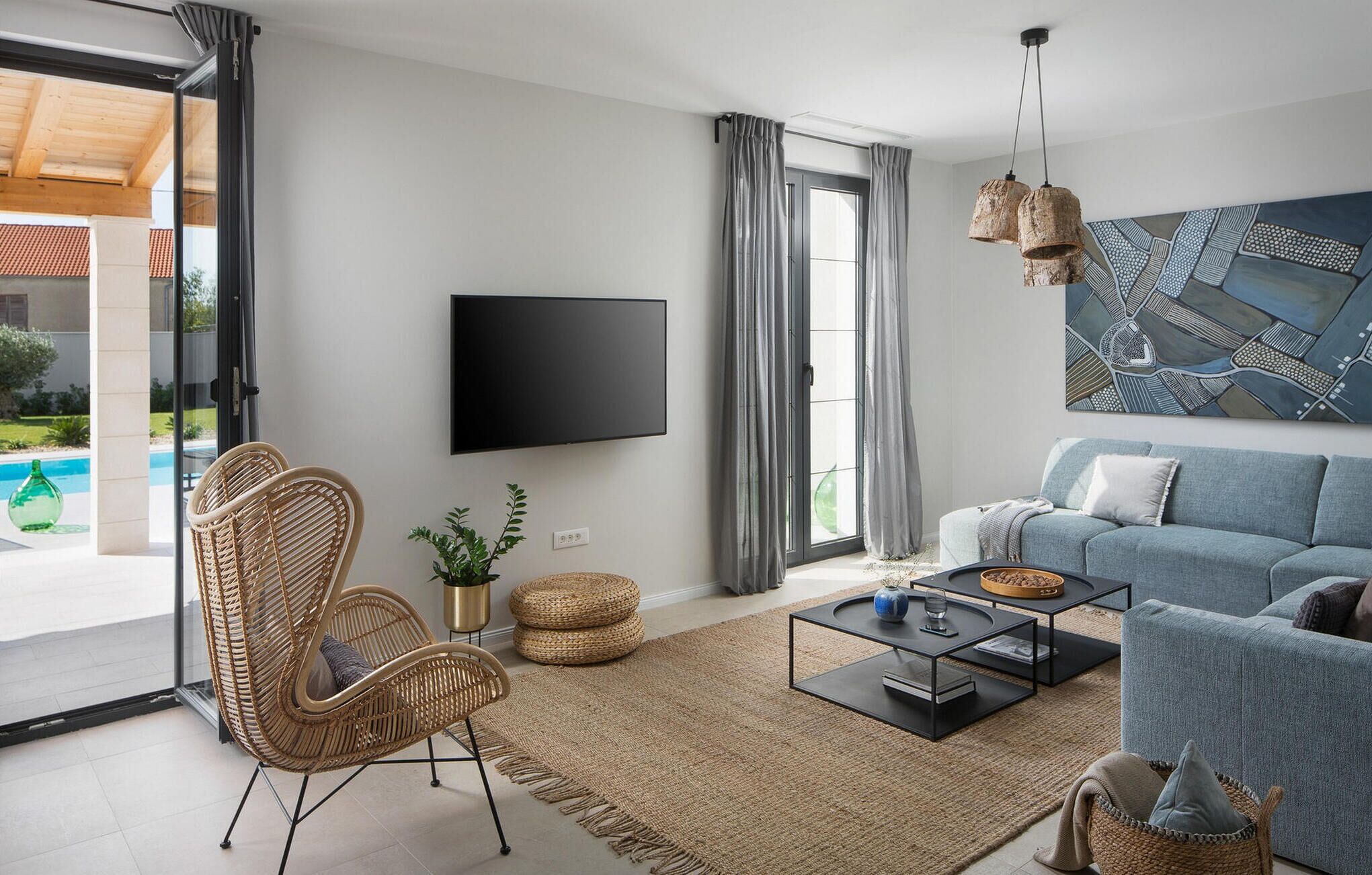 A modern living room with a flat-screen TV, a grey sofa, a wicker chair, a coffee table, and a large abstract painting.