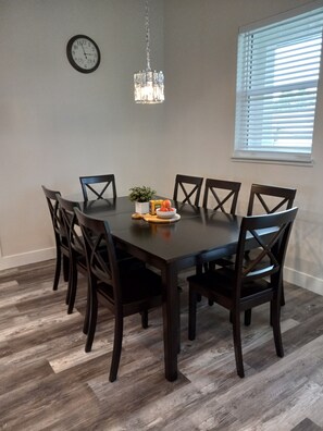 Un coin repas avec une table et des chaises en bois foncé, une horloge au mur et une fenêtre avec des stores.
