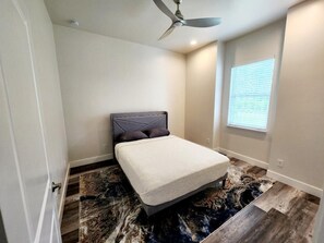Une chambre à coucher avec un lit, un ventilateur de plafond, une fenêtre avec des stores et un tapis à motifs.