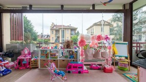 A child's playroom with toys, a pink stroller, and a large window with a view of buildings. - Beautiful 2-bed Apartment in Torino (Turin)