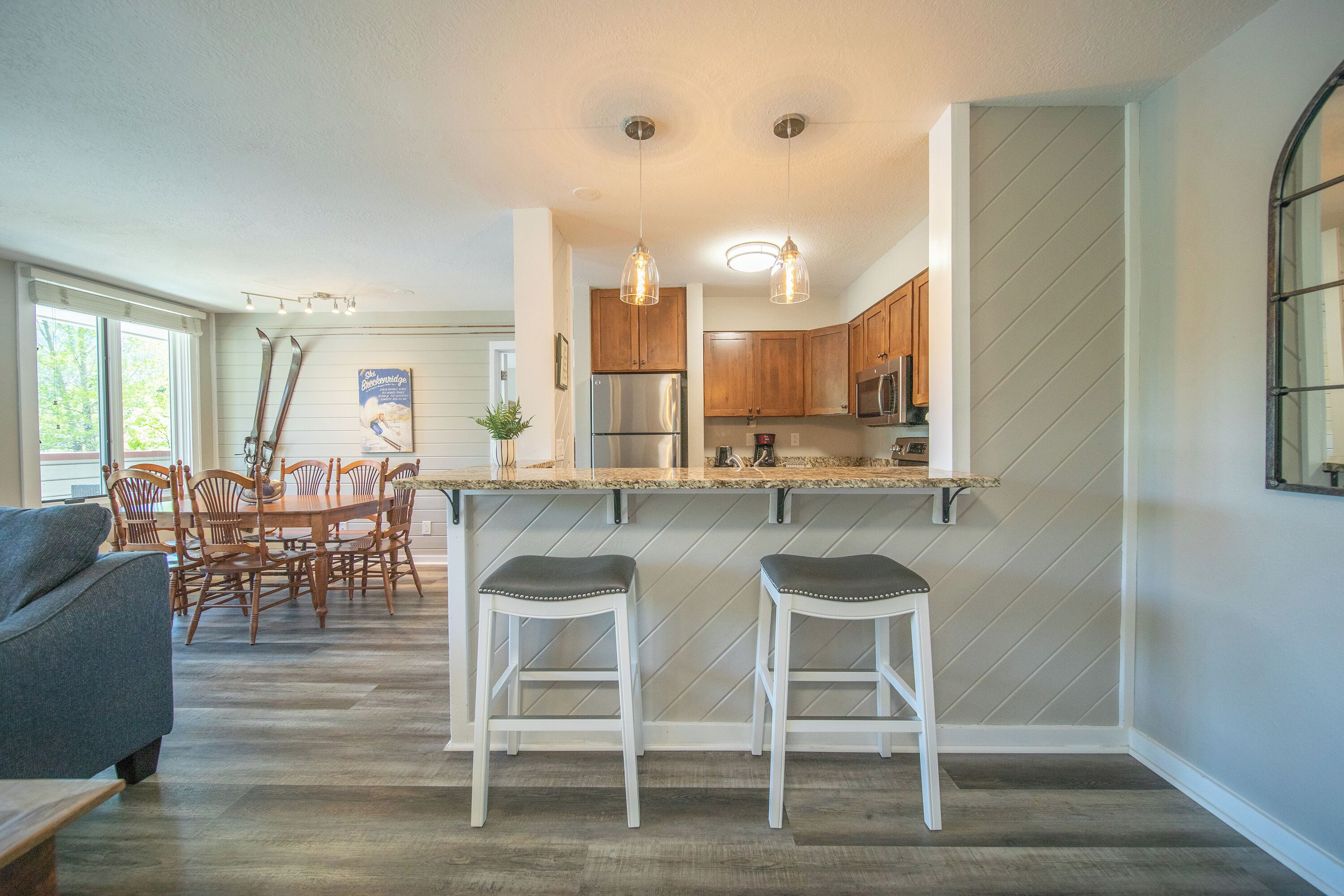 A modern kitchen with a bar area, wooden cabinets, stainless steel appliances, and a dining area with a gray sofa and wooden chairs.