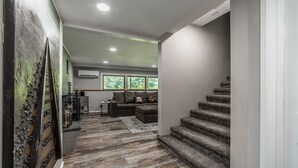 Een moderne woonkamer met een grijze bank, een trap en uitzicht op de omgeving door grote ramen.