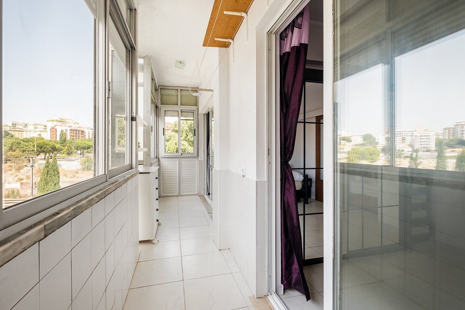 A narrow balcony with a view of buildings and trees, a sliding door, and a tiled floor.