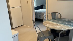 Uma cozinha compacta com geladeira, uma mesa de jantar com cadeiras e um quarto visível por uma porta aberta.