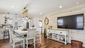 A modern kitchen with a dining area, wooden flooring, and a large flat-screen TV mounted on the wall.
