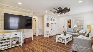 A living room with a flat-screen TV, wooden flooring, a ceiling fan, and a grey sofa.
