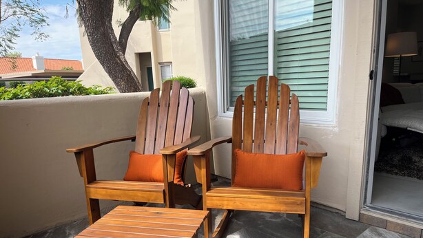 Deux fauteuils Adirondack en bois avec des coussins orange, une table en bois et une vue sur un bâtiment doté d’une fenêtre et de stores.