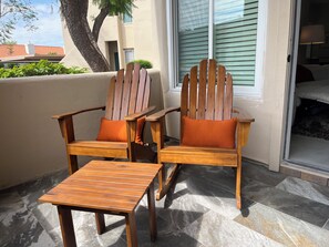 Two wooden Adirondack chairs with orange cushions, a wooden table, and a view of a building with a window and blinds.