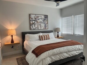 A bedroom with a bed, two bedside lamps, a ceiling fan, a window with blinds, and a painting on the wall.