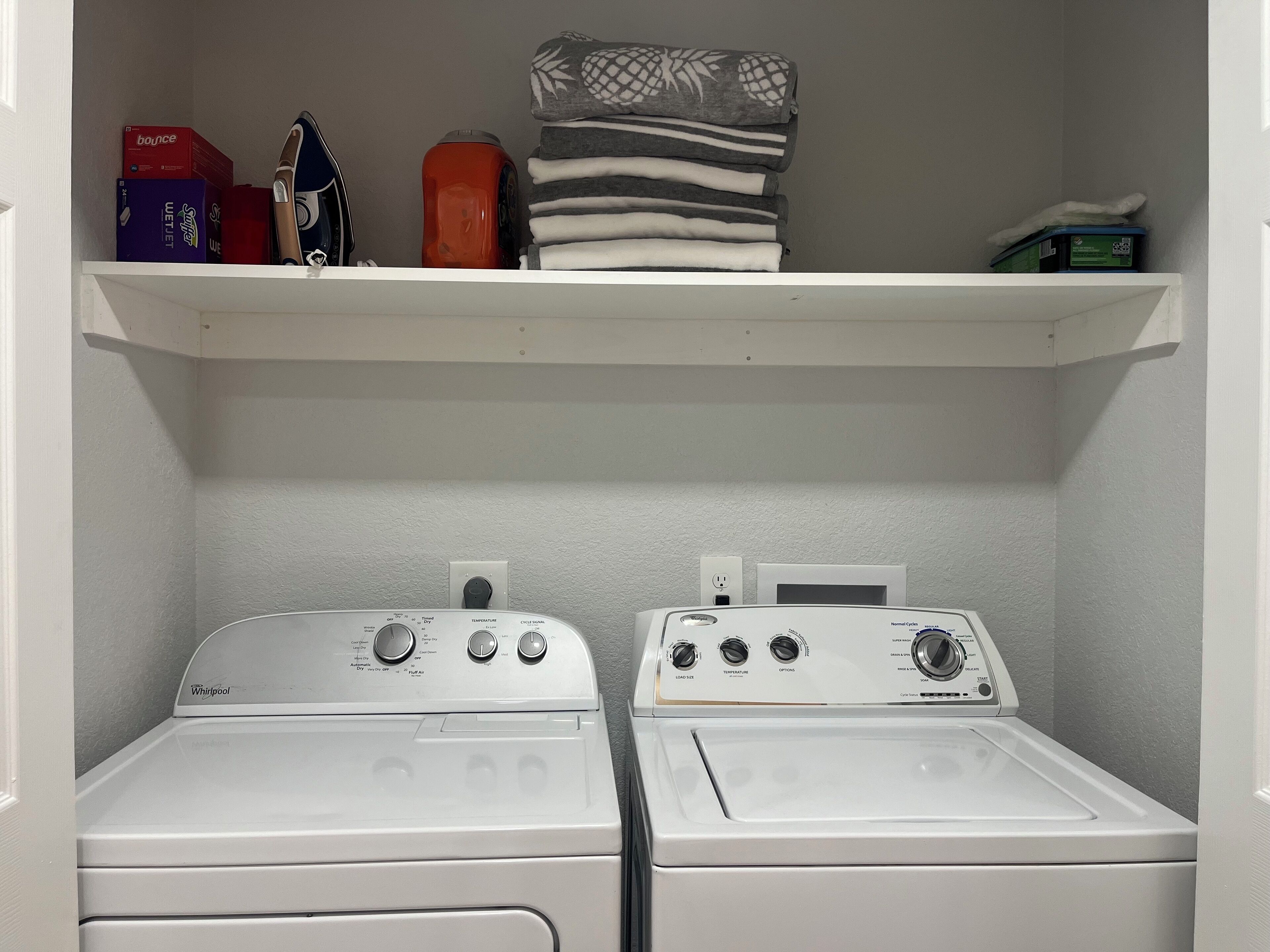 Een wasserette met twee witte Whirlpool wasmachines en drogers, een plank erboven met opgevouwen handdoeken en een doosje Bounce wasverzachter.