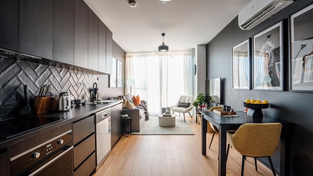 A modern kitchen with dark cabinetry, a herringbone backsplash, and a built-in stove. There is a dining area with a table and chairs, and a living room with a sofa and a coffee table. - Midnight Moonshine Loft ITAS (LAUNCESTON)