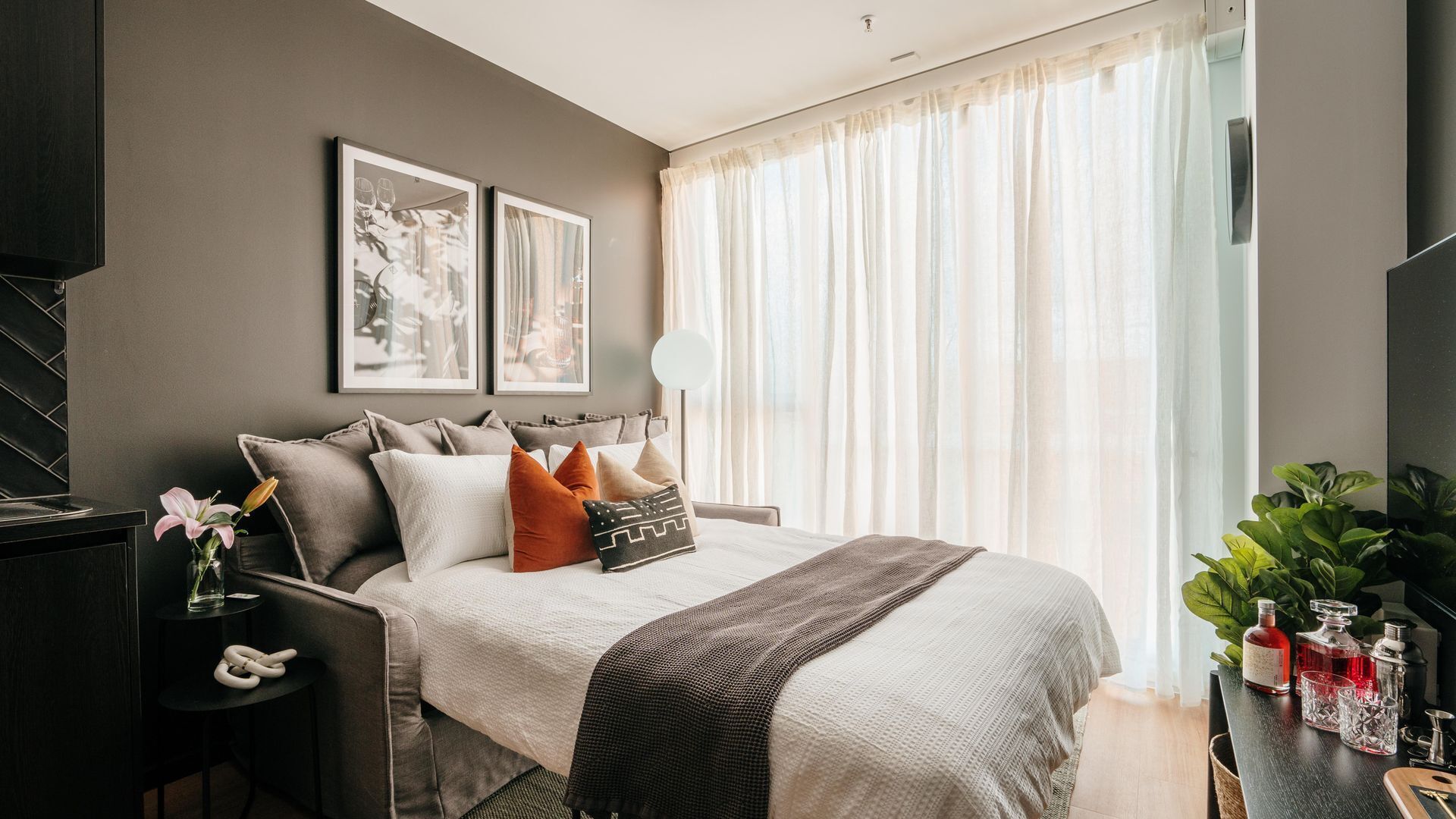 Una habitación de hotel moderna con una cama grande, un escritorio con silla, una mesita auxiliar y un amplio ventanal con cortinas translúcidas.