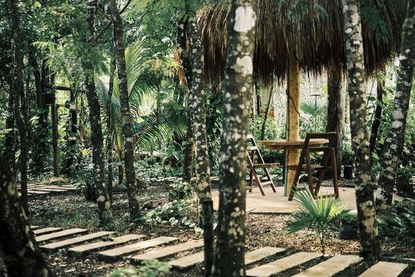 A wooden table and chairs under a thatched roof in a lush garden with a stone pathway.