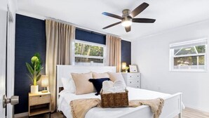 A modern bedroom with a large bed, a ceiling fan, a bedside table with a lamp, a window with blinds, and a potted plant. - Free Heated Pool Oasis: Coastal Bungalow Near Town & Beach (Bradenton)