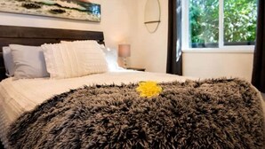 A bedroom with a bed, pillows, a fluffy rug, a painting, a mirror, and a window with a view of trees.