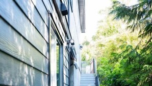 A wooden deck leading to a house with a window and a door, surrounded by greenery.