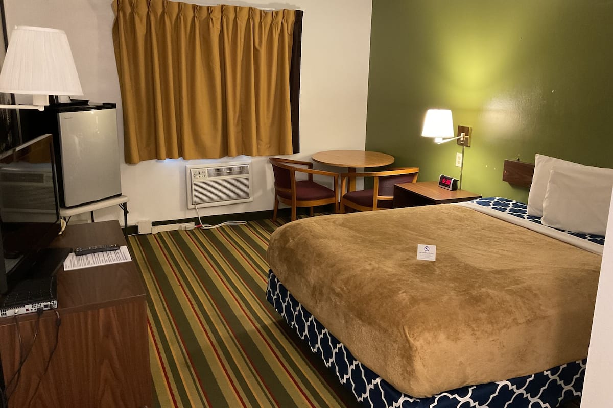 A hotel room with a bed, a desk, a chair, a lamp, a clock, and a television.