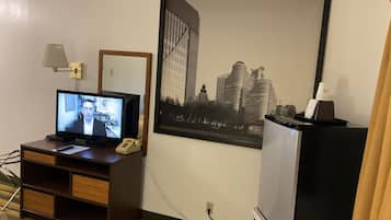 Quarto de hotel com televisão, escrivaninha com luminária, telefone, quadro com vista urbana e minibar.