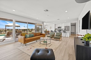 A spacious living area with a sofa, ottoman, and chairs, a kitchen in the background, and a large window overlooking a pool. - Stunning Desert Getaway w/ Ice Bath & Pool! (Bermuda Dunes)