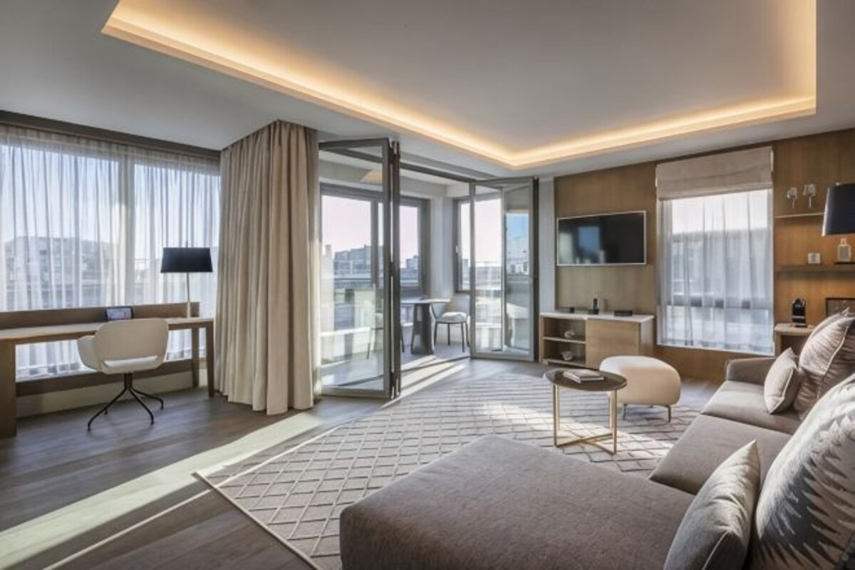 A modern hotel room with a sofa, a desk, a chair, a TV, and a balcony with a city view.